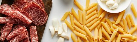 Panoramic shot of meat platter, garlic, pasta and bowl with mozzarella on white background