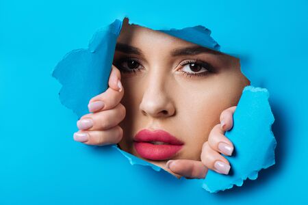 Beautiful woman with smoky eyes and pink lips looking at camera across hole and touching blue paper