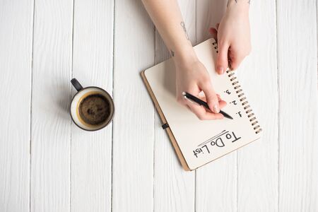 Cropped view of woman writing in notebook with to do list lettering near cup of coffee on wooden...