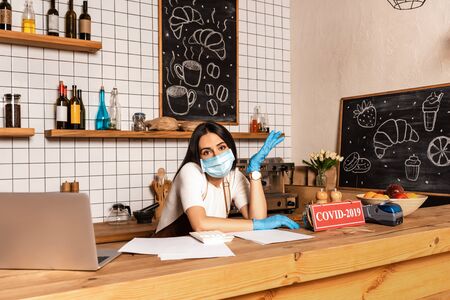Cafe owner looking at camera near table with laptop, papers, card with covid-2019 lettering, payment terminal and bowl with fruitsの写真素材