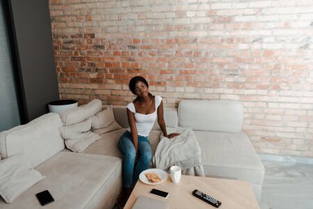 High angle view of african american woman on sofa near coffee table with plate, cup of tea, smartphone and remote controller in living roomの写真素材