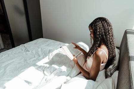 African american woman reading book on bed in bedroomの写真素材