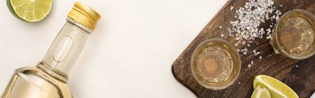 top view of golden tequila with lime, salt on wooden cutting board on white marble surface,...