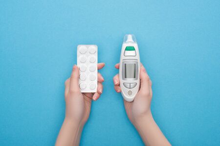 partial view of woman holding pills in blister pack and ear thermometer on blue background