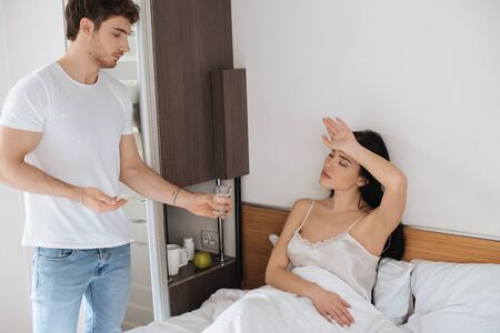 boyfriend giving water and pill to tired sick girlfriend with headache in bedroom during self isolationの写真素材