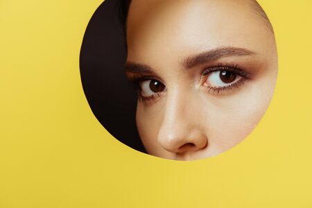 Girl with smoky eyes looking at camera across hole in yellow paper on black backgroundの写真素材