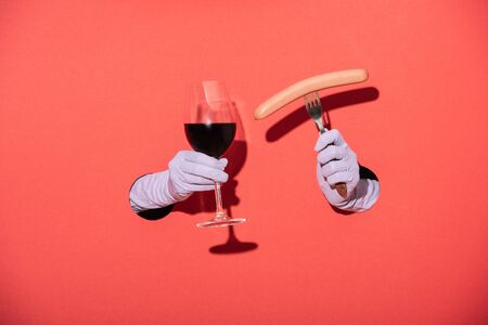 cropped view of woman in gloves holding glass with red wine and fork with sausage on redの写真素材