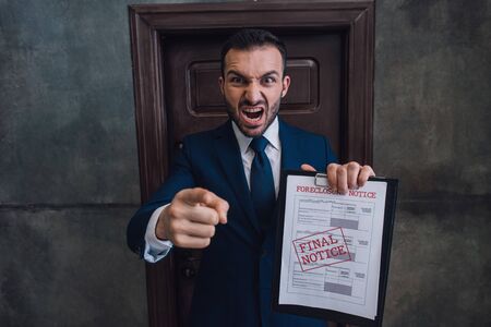 Angry collector with documents with foreclosure lettering pointing with finger and shouting in room