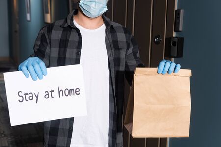 Cropped view of courier in medical mask holding package and card with stay at home lettering on...