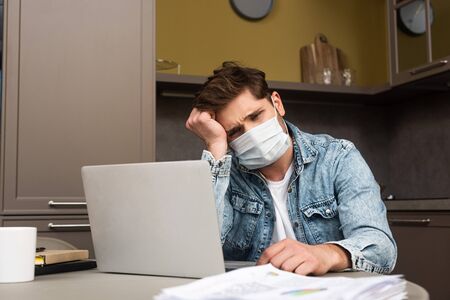 Selective focus of sad freelancer in medical mask sitting at table with papers and laptop