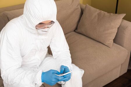 Man in latex gloves, medical mask and hazmat suit using smartphone on couch at home