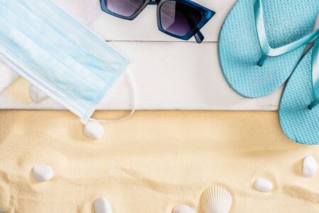 Top view of medical mask, flip flops and sunglasses on white wooden planks near pebbles on sand