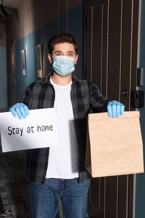 Courier in medical mask and latex gloves holding card with stay at home lettering and package on porchの写真素材