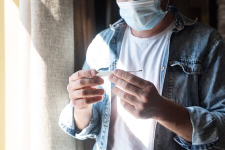 Cropped view of man in medical mask holding thermometer near window at home