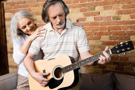 bearded and mature man playing acoustic guitar near smiling wife