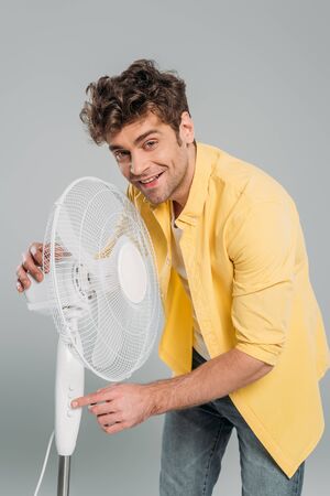 Man turning on electric fan, smiling and looking at camera isolated on greyの写真素材