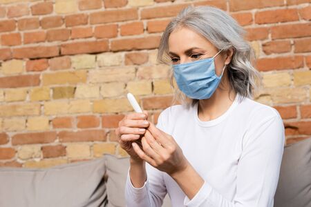 sick and mature woman in medical mask looking at digital thermometer