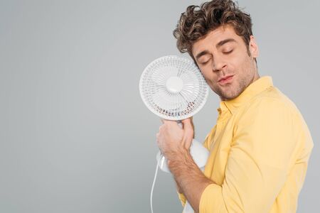 Man with closed eyes holding desk fan isolated on grey