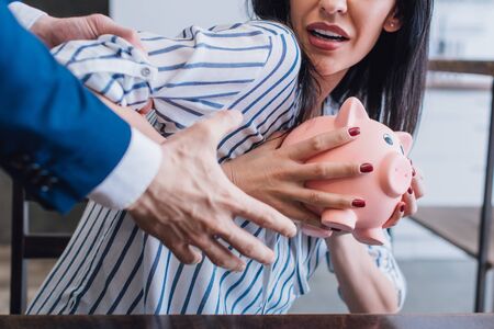 Cropped view of collector grabbing female hand and scared woman holding piggy bank at table in room