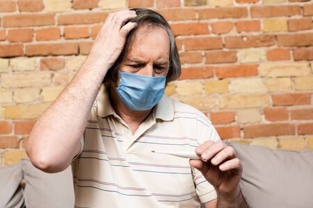 mature man in medical mask looking at digital thermometer