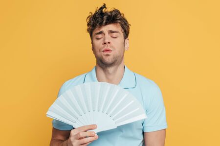 Man with closed eyes and hand fan suffering from heat isolated on yellowの写真素材