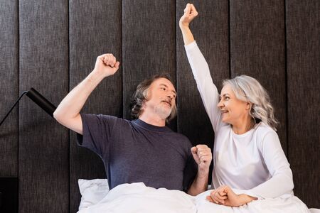 happy mature man and woman with hands above head looking at each other in bedroomの写真素材