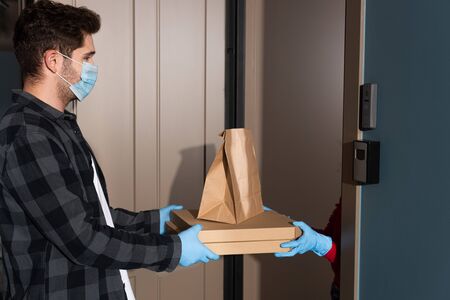 Side view of courier in medical mask giving package and pizza boxes to woman near open doorの写真素材