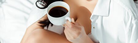 Cropped view of sexy woman in shirt and bra holding coffee on bed, panoramic shotの写真素材