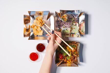 top view of woman holding chopsticks near takeaway boxes with prepared chinese food and sauces on...