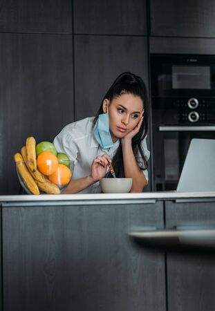 pensive woman holding spoon while looking at laptop near bowl with fruits in kitchenの写真素材
