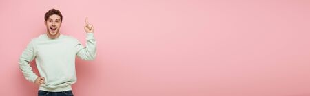 panoramic crop of excited guy showing idea sign while standing with hand on hip and looking at camera  on pink backgroundの写真素材