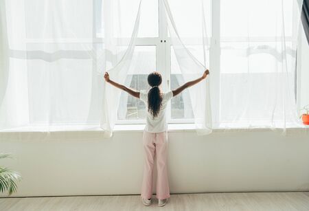 back view of african american girl in pajamas opening window curtains in sunlightの写真素材