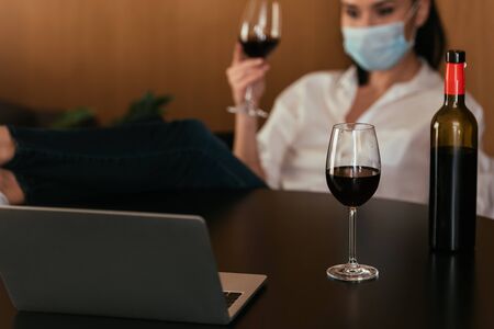 selective focus of woman in protective mask holding glass of red wine during video chat on laptop