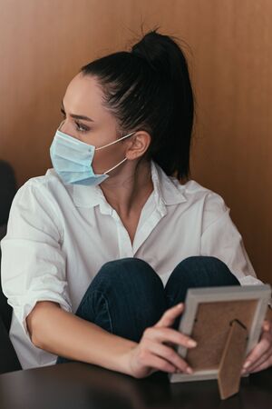 depressed young woman in medical mask looking away while holding photo frameの写真素材