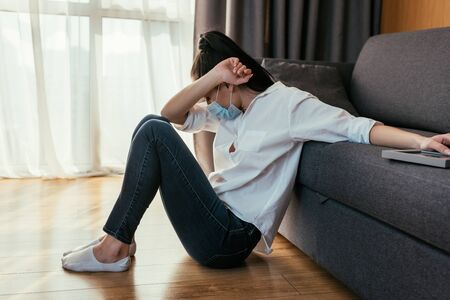 grieving young woman in medical mask obscuring face with hand while sitting on floor and touching photo frameの写真素材