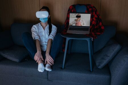 young woman sitting on sofa near laptop with boyfriend eating popcorn while using vr headsets...