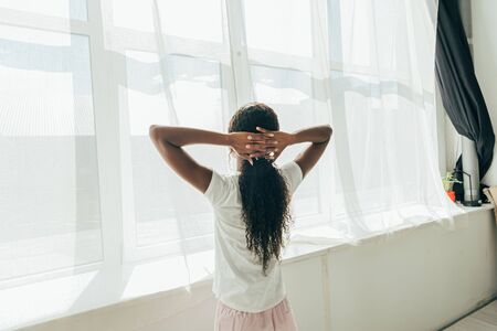 back view of african american girl standing by window in sunshine with hands behind headの写真素材