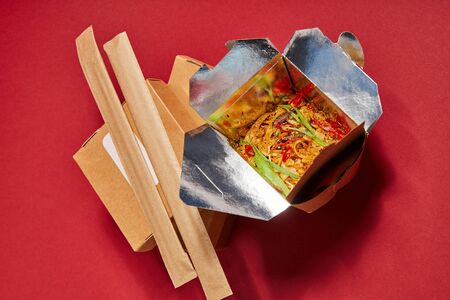 top view of chopsticks in paper packaging near tasty noodles in takeaway box on redの写真素材