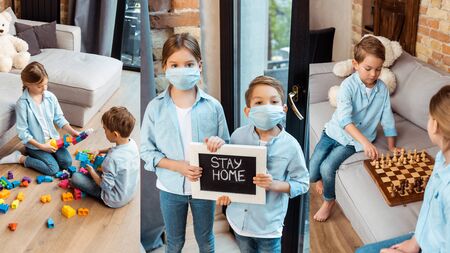 collage of siblings in medical masks holding chalkboard with stay home lettering, playing chess and building blocks at homeの写真素材