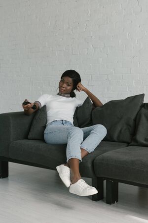 pretty african american girl holding tv remote controller while sitting on sofa at homeの写真素材