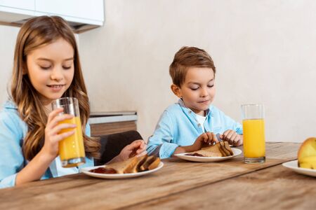 selective focus of happy sister holding glass of orange juice near brother and tasty breakfastの写真素材