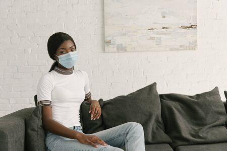 pensive african american girl in medical mask sitting on grey sofa at home