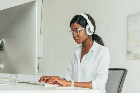 attentive african american freelancer in wireless headphones and glasses typing on computer keyboard