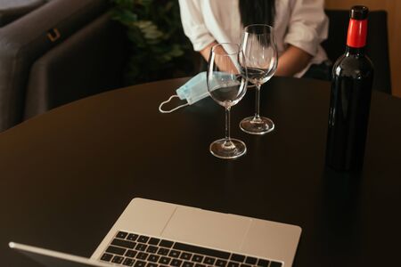 cropped view of woman sitting at table near wine glasses, bottle, medical mask and laptopの写真素材