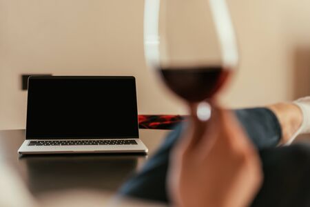 selective focus of woman holding glass of red wine near laptop with blank screen
