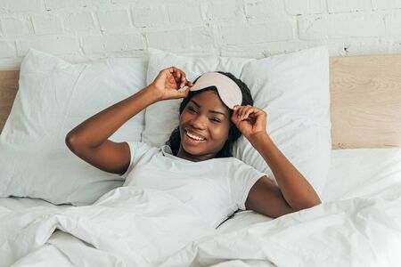 smiling african american girl touching sleep mask while lying in bed and looking at cameraの写真素材