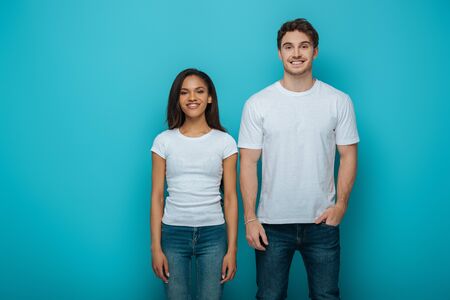 happy man with hand in pocket and cheerful african american girl smiling at camera on blue backgroundの写真素材