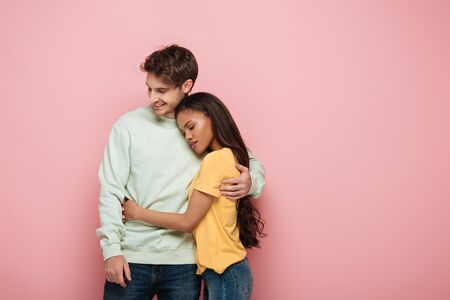 attractive african american girl with closed eyes and happy guy embracing on pink background