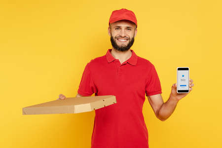 KYIV, UKRAINE - MARCH 30, 2020: smiling bearded delivery man in red uniform with pizza box showing smartphone with messenger app on yellowのeditorial素材