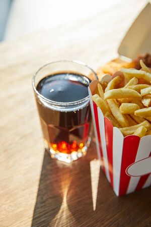 selective focus of french fries and soda in glass on wooden table in sunlightの写真素材